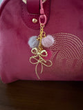 Handmade pink and grey fuzzy pom-pom bag charm with gold wire bows, shown attached to a pink velvet purse.