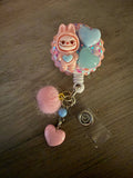 Cute badge reel featuring a pink monster character, two hearts, a pink pom-pom, and colorful charms.