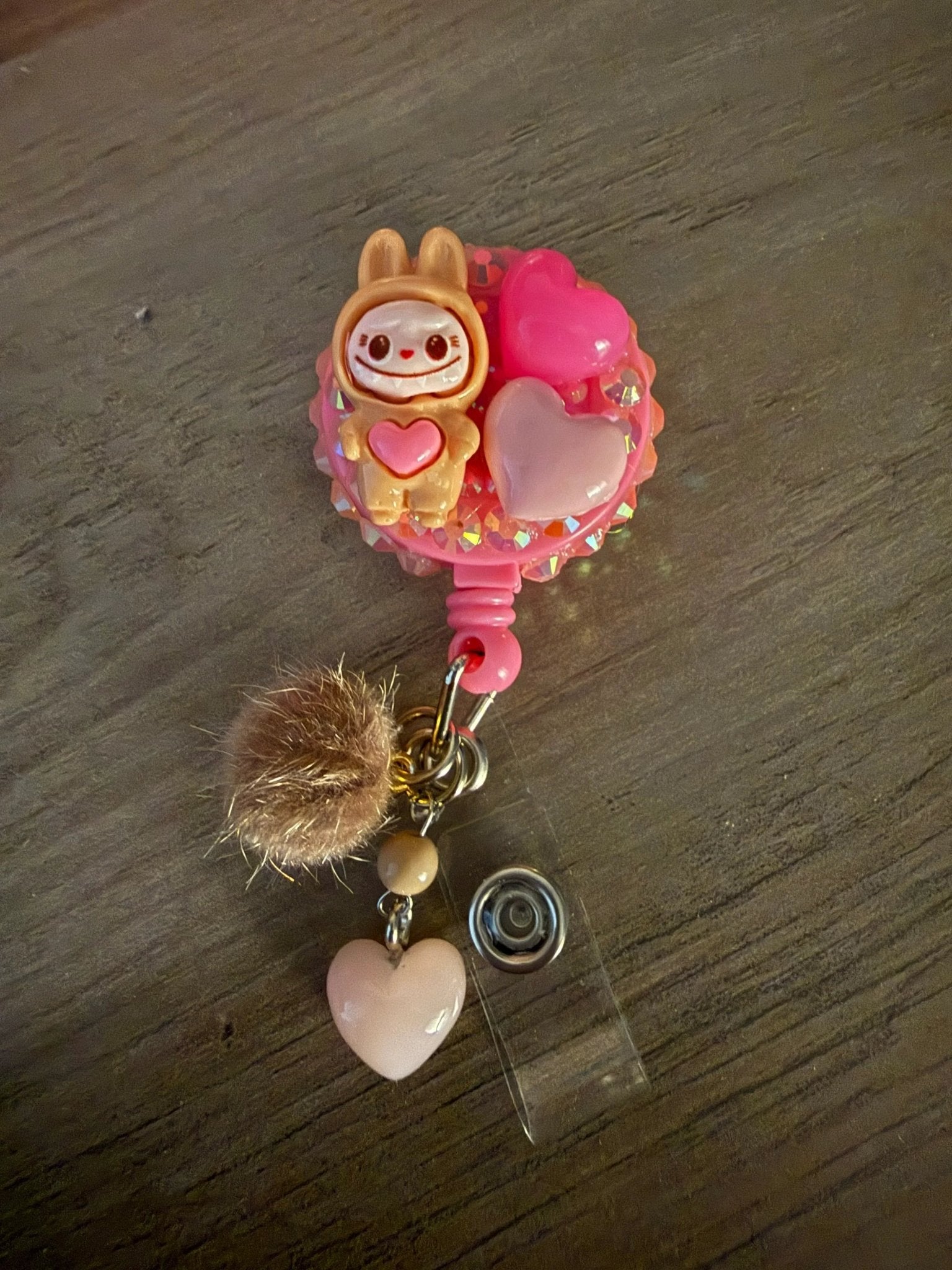 Cute badge reel featuring a tan monster character in a hood, two colorful hearts, a fluffy pom-pom, and a pink heart charm.