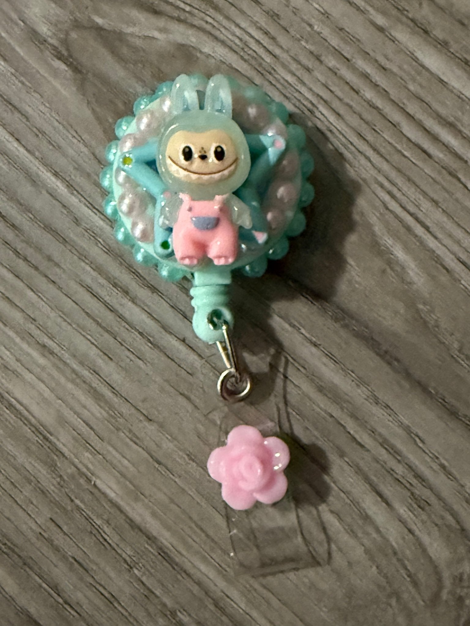 Handmade teal decoden badge reel featuring a Labubu character in pink overalls, white pearls, and a pink rose charm.