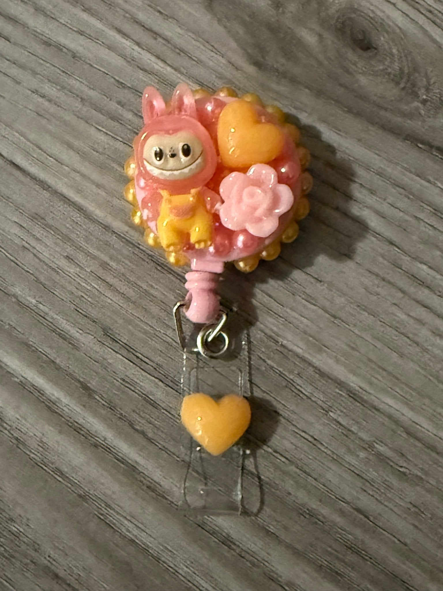 Handmade pink and yellow decoden badge reel featuring a Labubu character, an orange heart, and a pink rose charm.