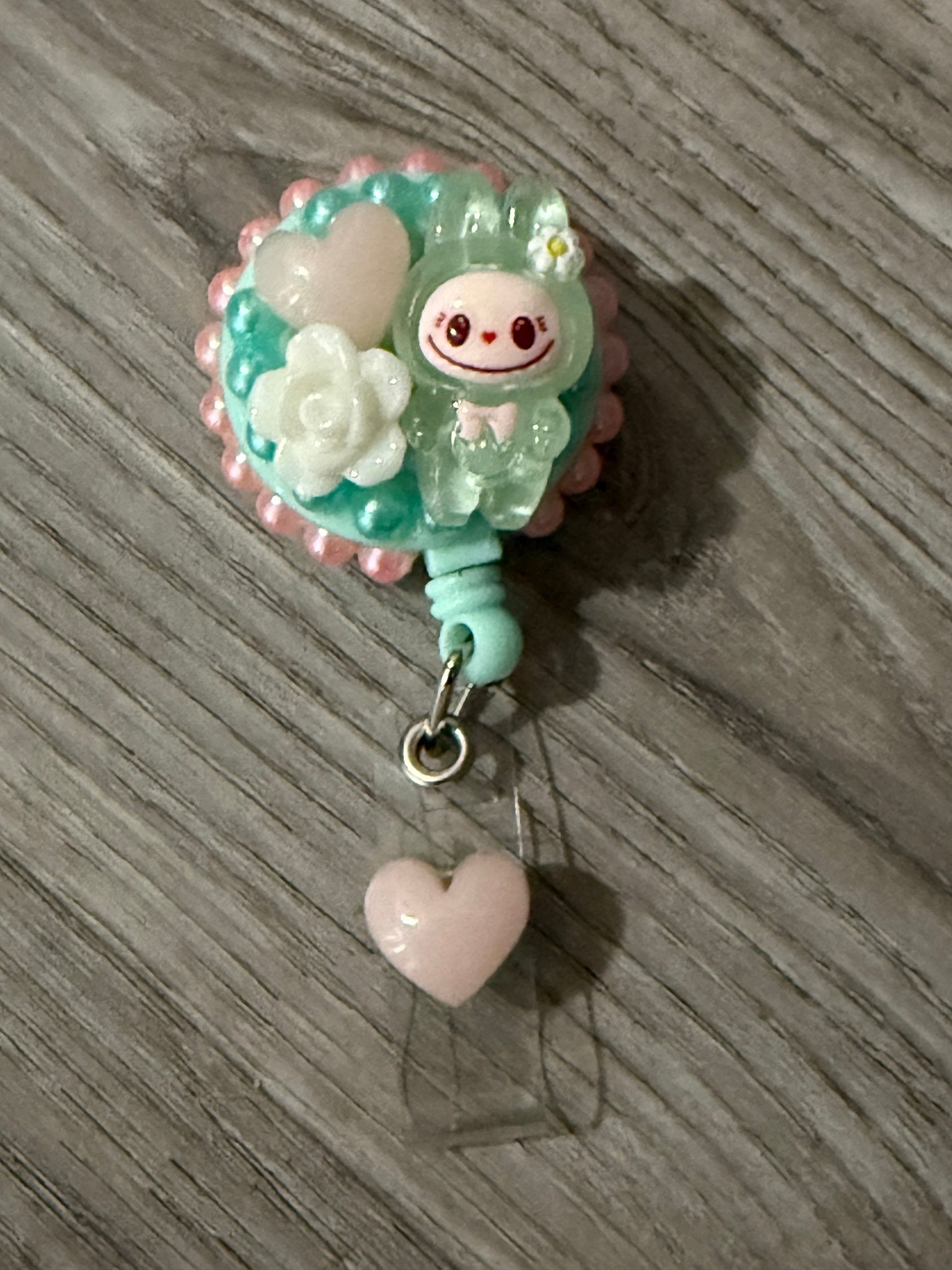 Handmade teal decoden badge reel featuring a green Labubu character, a white rose charm, and a pale pink heart.