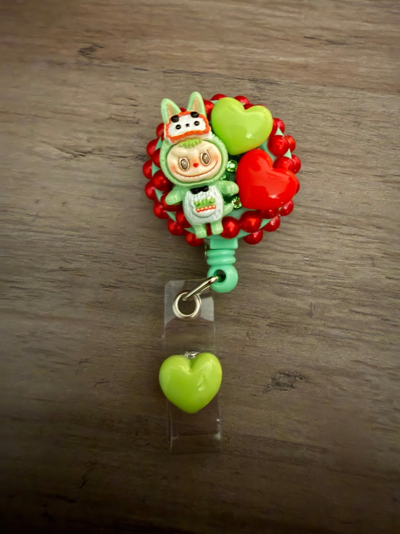 Decoden retractable badge reel featuring a green character with lime and red heart charms on a red beaded base.