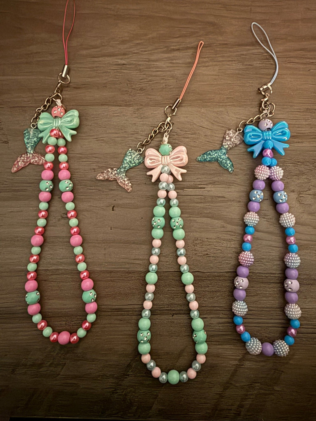 Three handmade beaded phone wristlets with mermaid tail charms and bows.