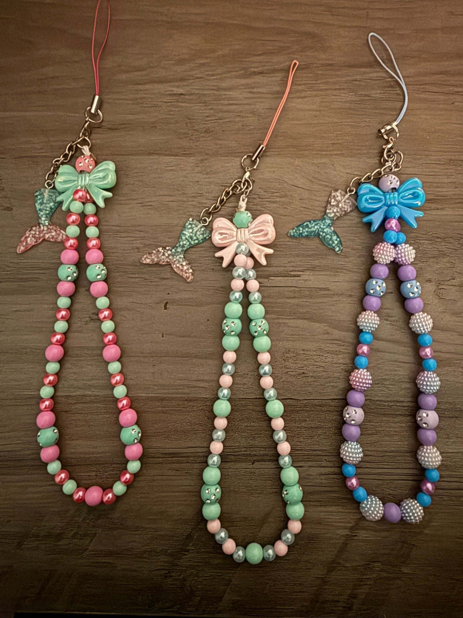 Three handmade beaded phone wristlets with mermaid tail charms and bows.