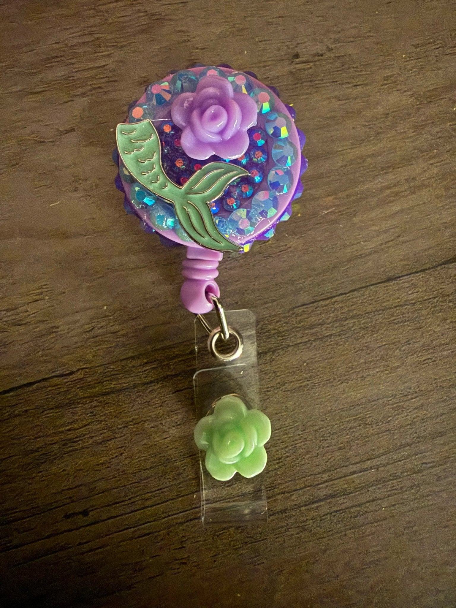 A purple badge reel with a mermaid tail design, a purple rose, and a small green flower charm attached.