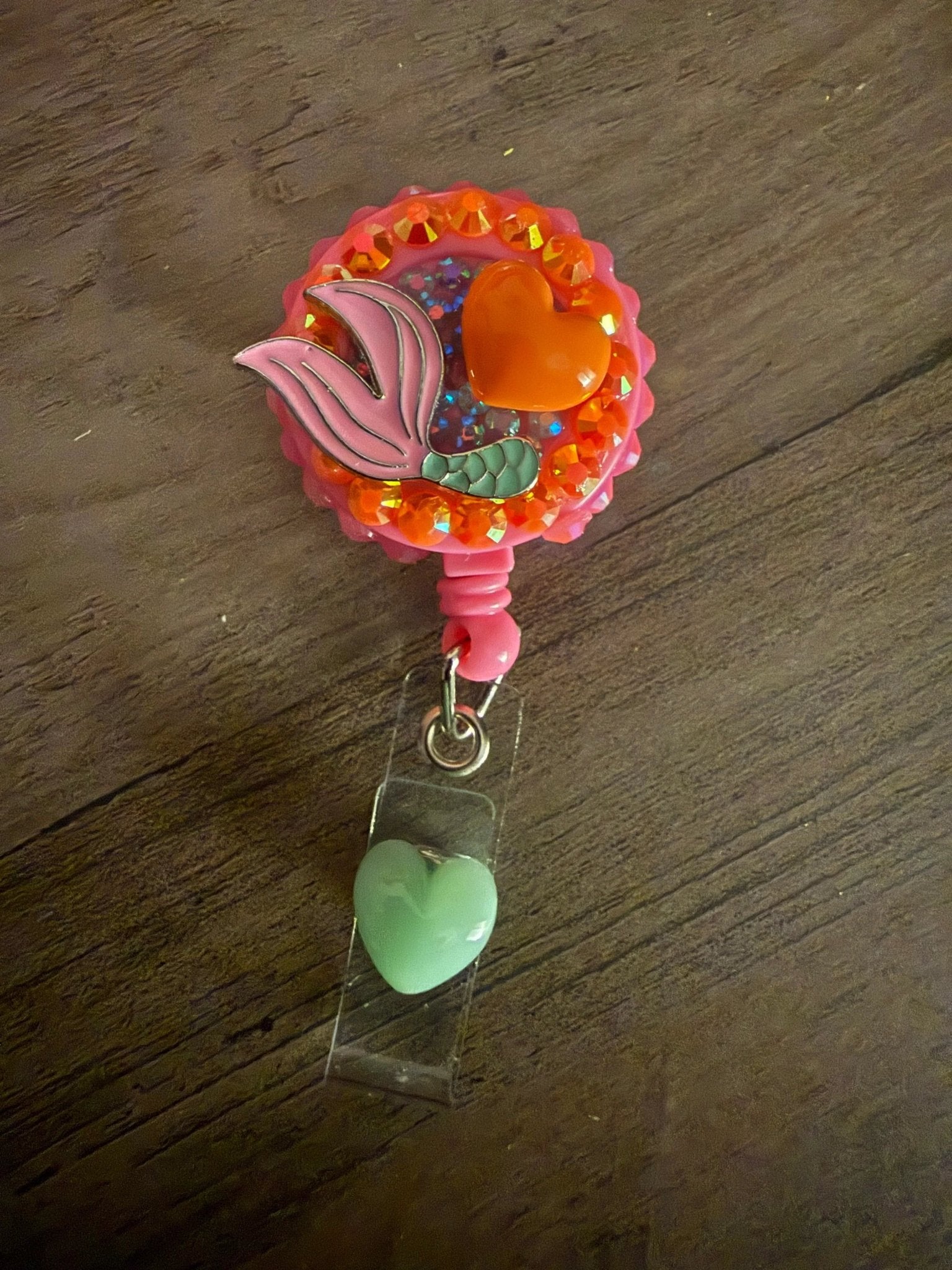 A pink badge reel featuring a mermaid tail design, an orange heart, and a green heart charm attached.