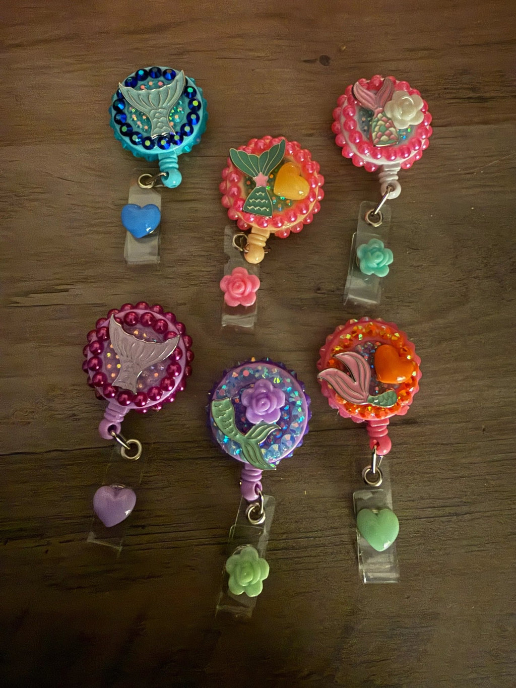 A collection of six colorful badge reels with mermaid and flower designs, with beads and small charms in various colors.