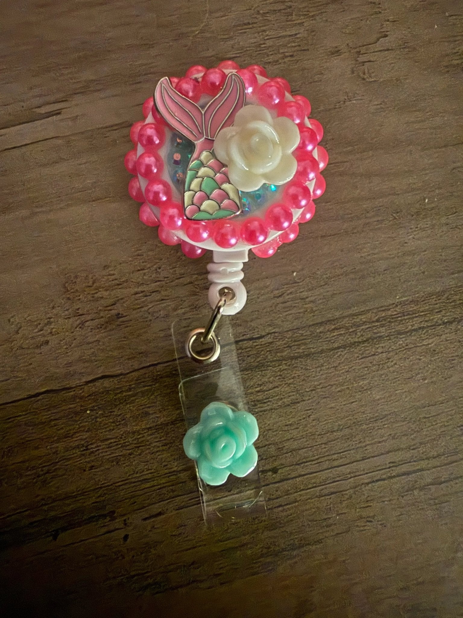 A pink badge reel with a mermaid tail design, a white flower, and a small turquoise rose charm attached.