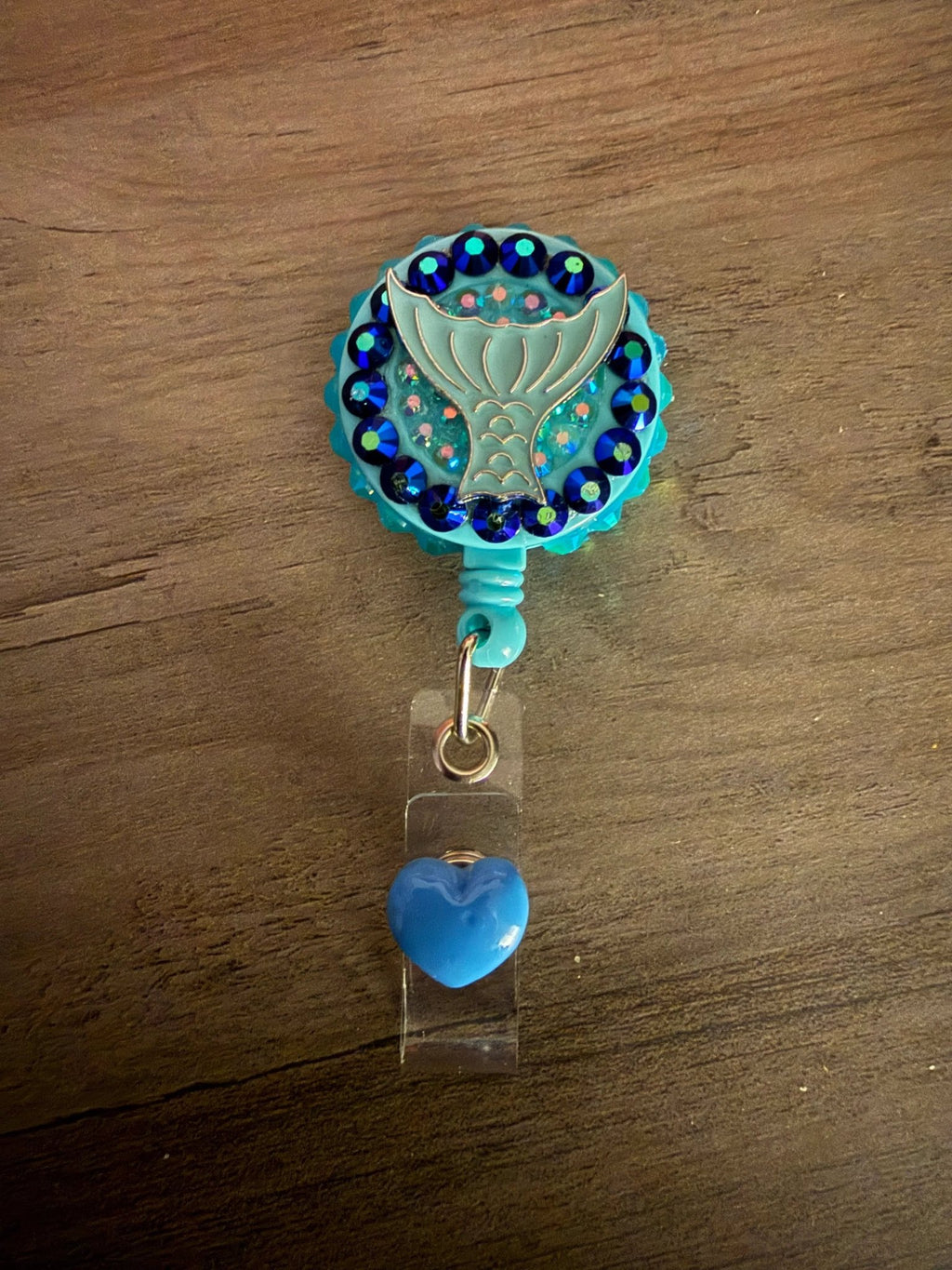 A turquoise badge reel featuring a mermaid tail design, surrounded by blue sequins, with a blue heart charm attached.