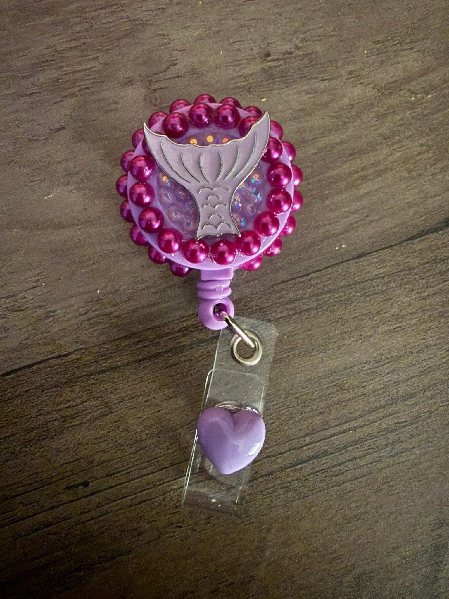 A purple badge reel with a mermaid tail design, surrounded by pink beads, and a small purple heart charm attached.