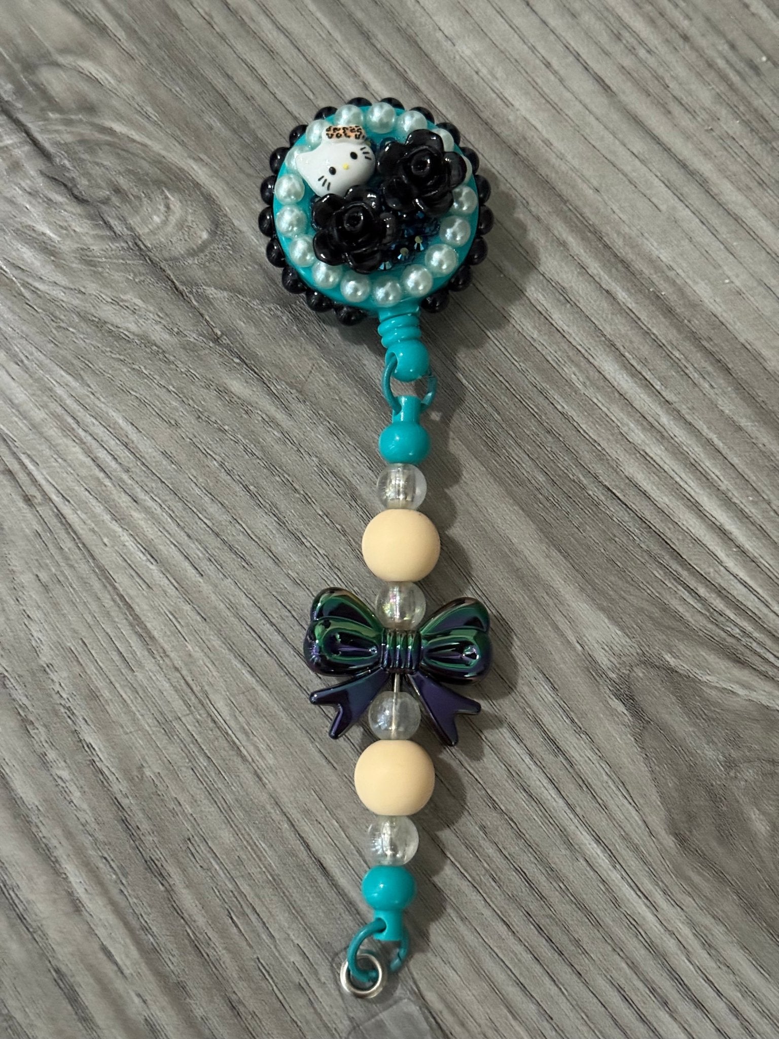 A colorful badge reel with a cute cat design and black roses, adorned with turquoise and pearl beads, and a decorative bow.