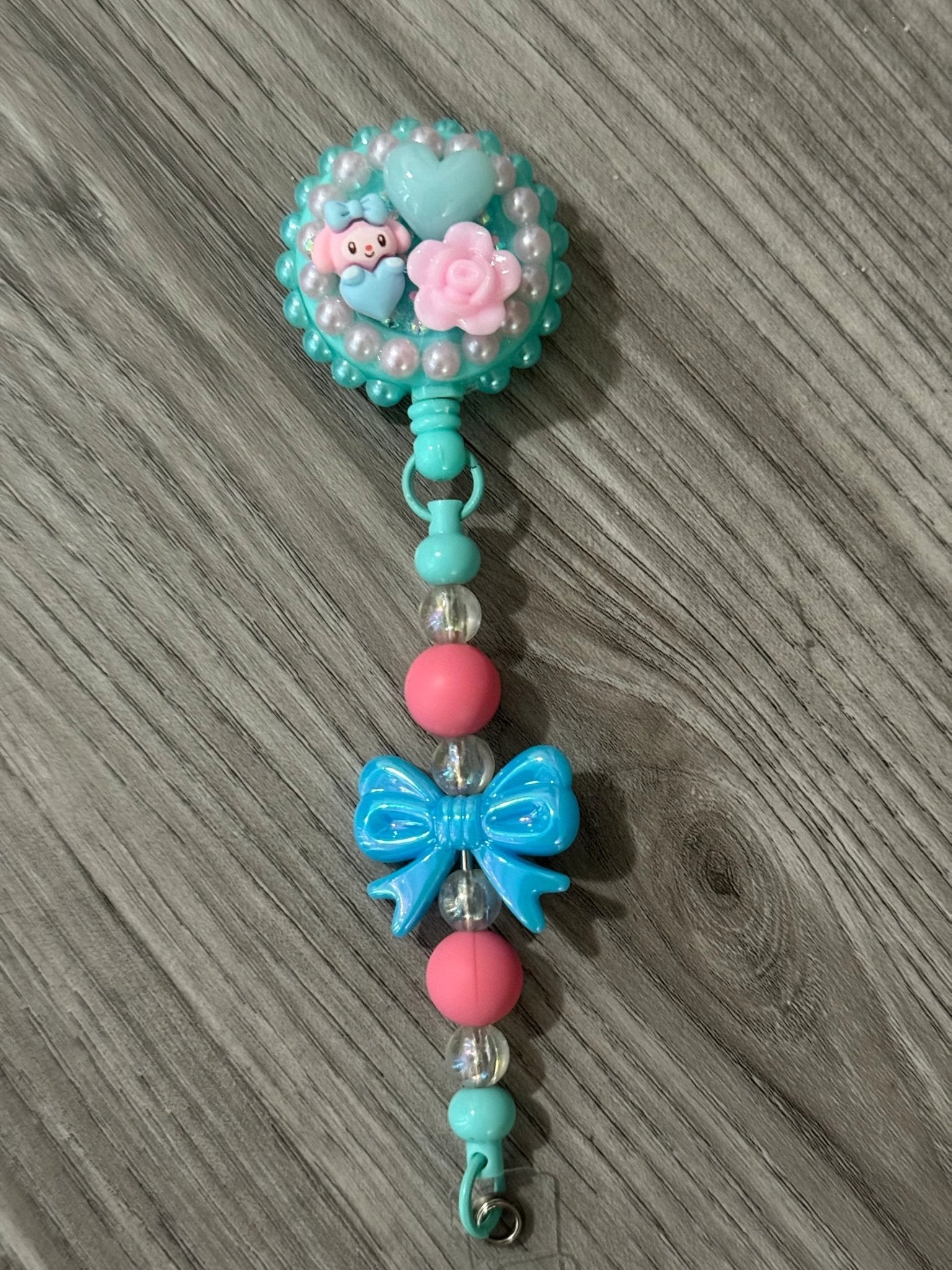 A turquoise badge reel with a cute character, pink flowers, and hearts, beaded strap with a blue bow and pink beads.