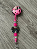A pink badge reel adorned with playful embellishments, including a heart, flowers, and cute characters, with colorful beads.