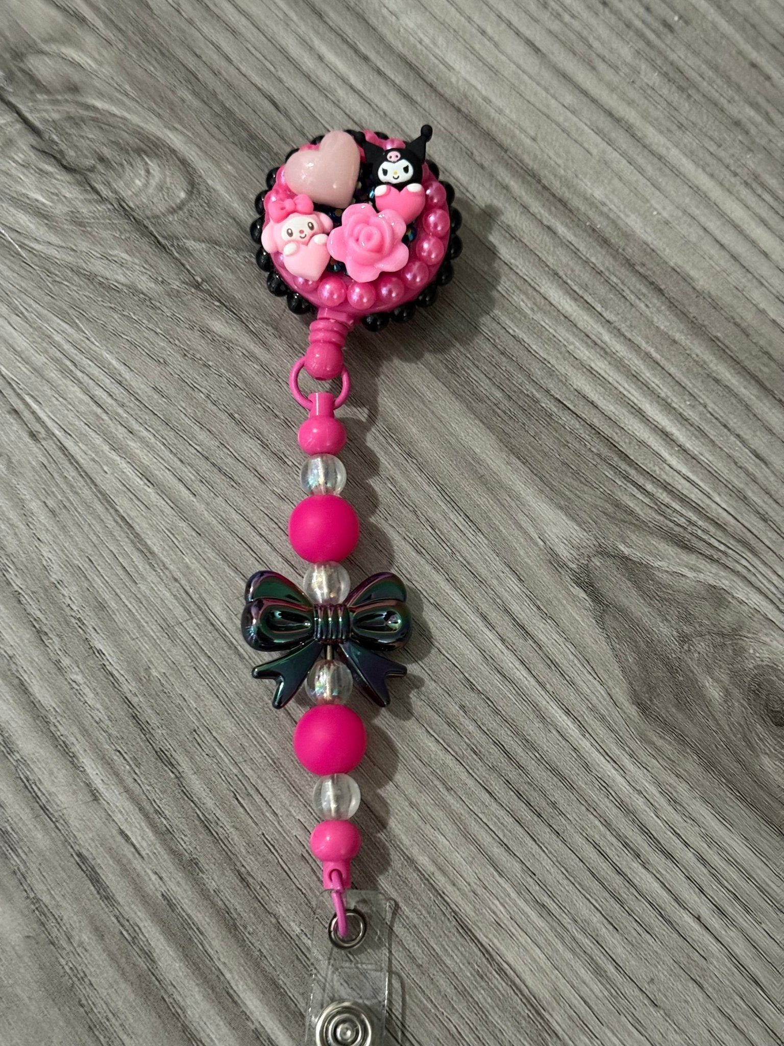 A pink badge reel adorned with playful embellishments, including a heart, flowers, and cute characters, with colorful beads.