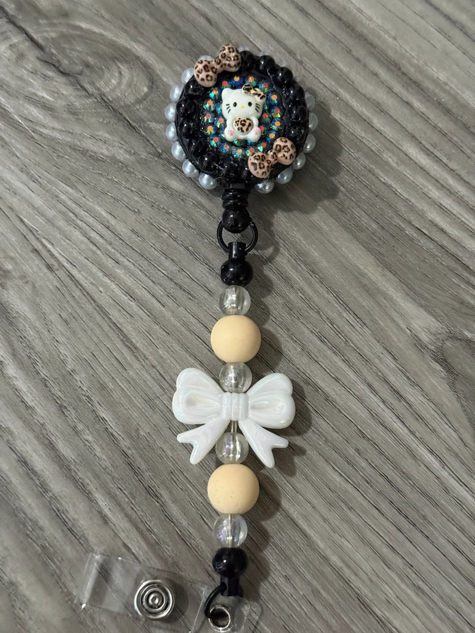 A whimsical badge reel with a cute cat design and leopard print bows, connected to a beaded strap with a white bow.