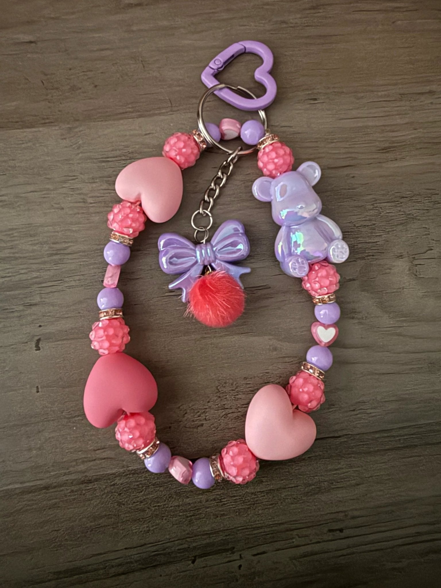 Colorful bag charms or phone charms with pink and purple heart beads, a bear figure, a bow charm, and a fluffy pom-pom.