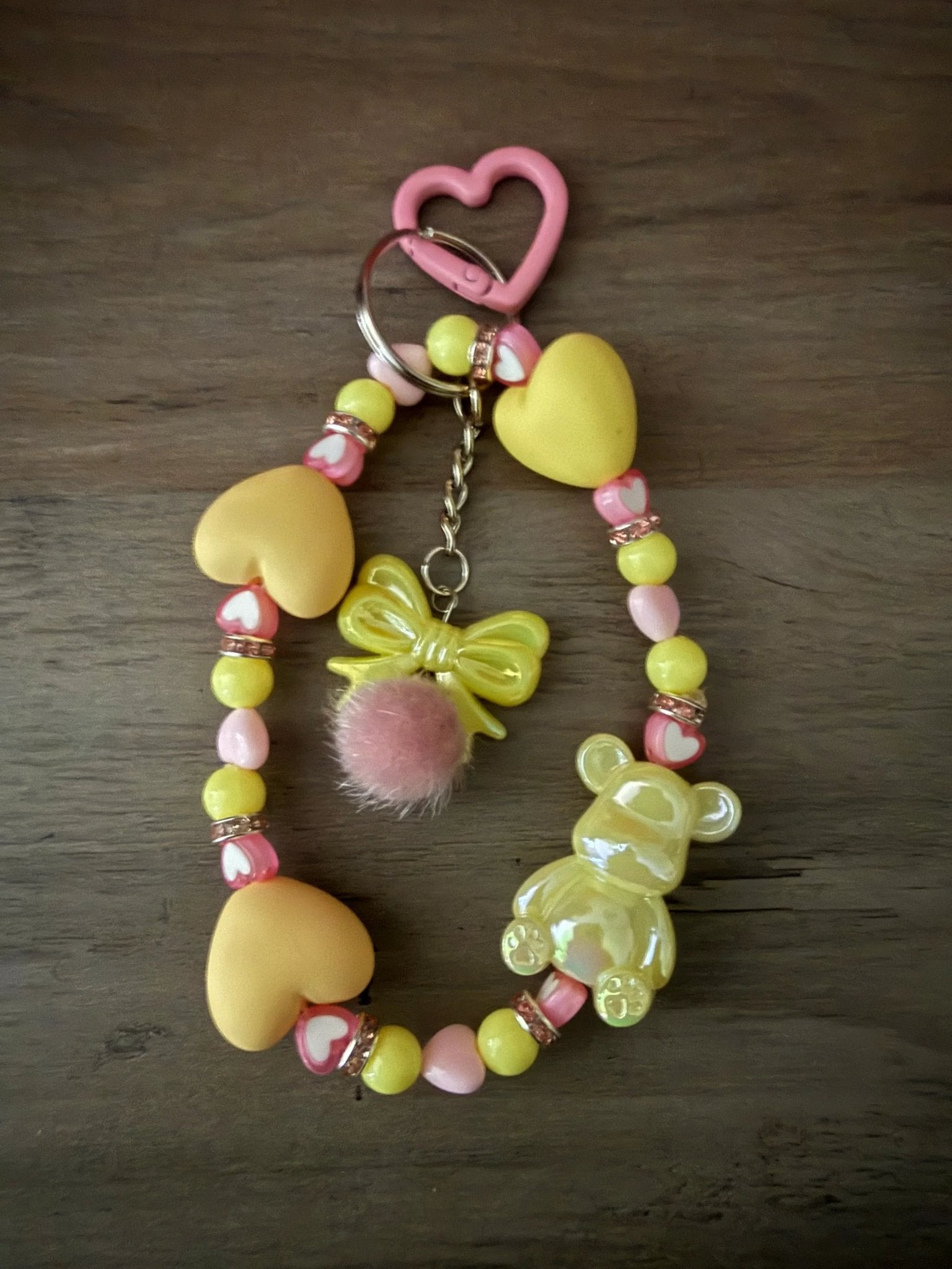 A yellow and pink bag charms or phone charms with heart beads, a fluffy pom-pom, a bow charm, and a bear figure.
