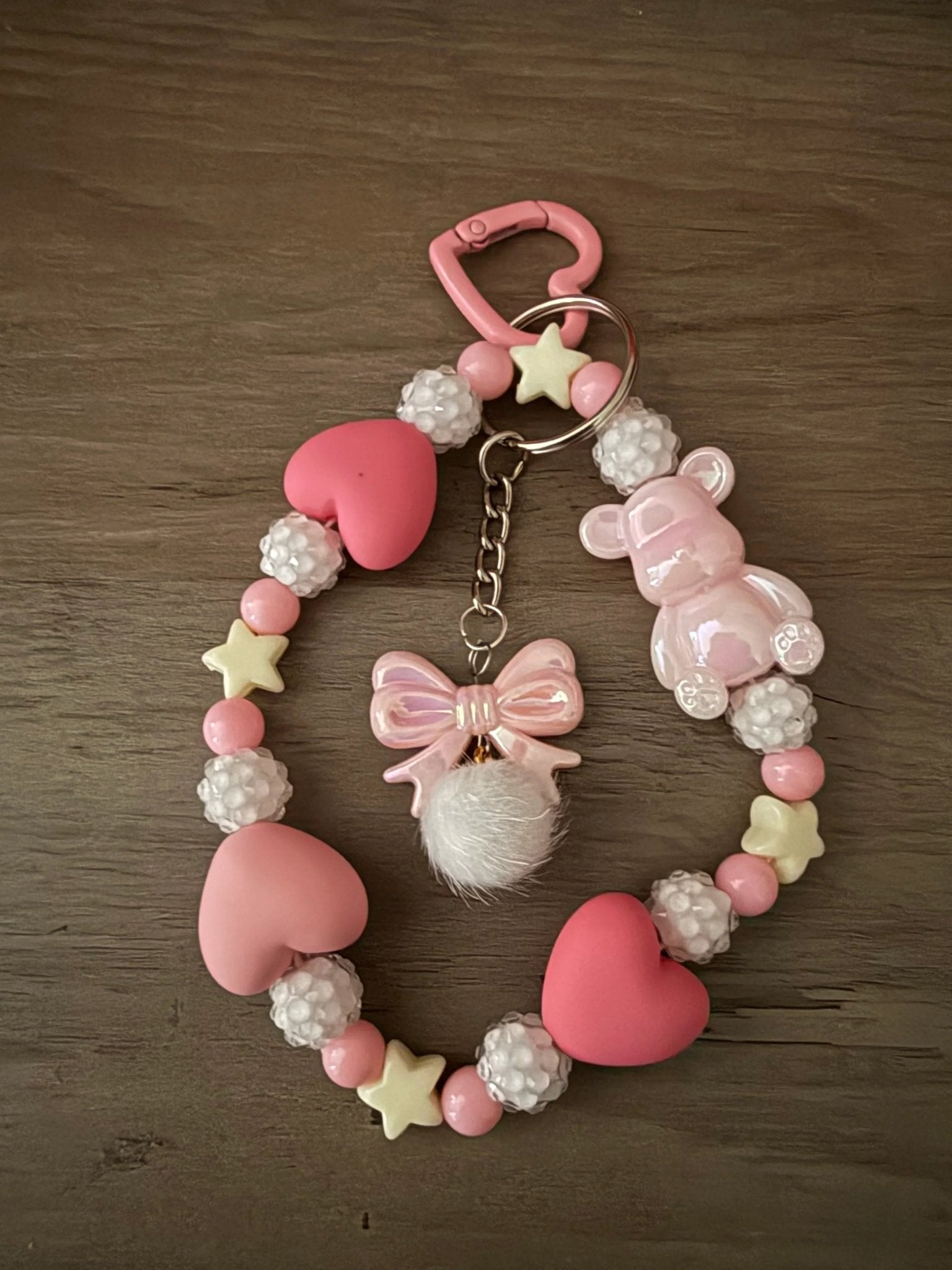 A pink bag charms or phone charms with heart-shaped beads, a cute bear figure, a bow charm, and fluffy pom-poms.