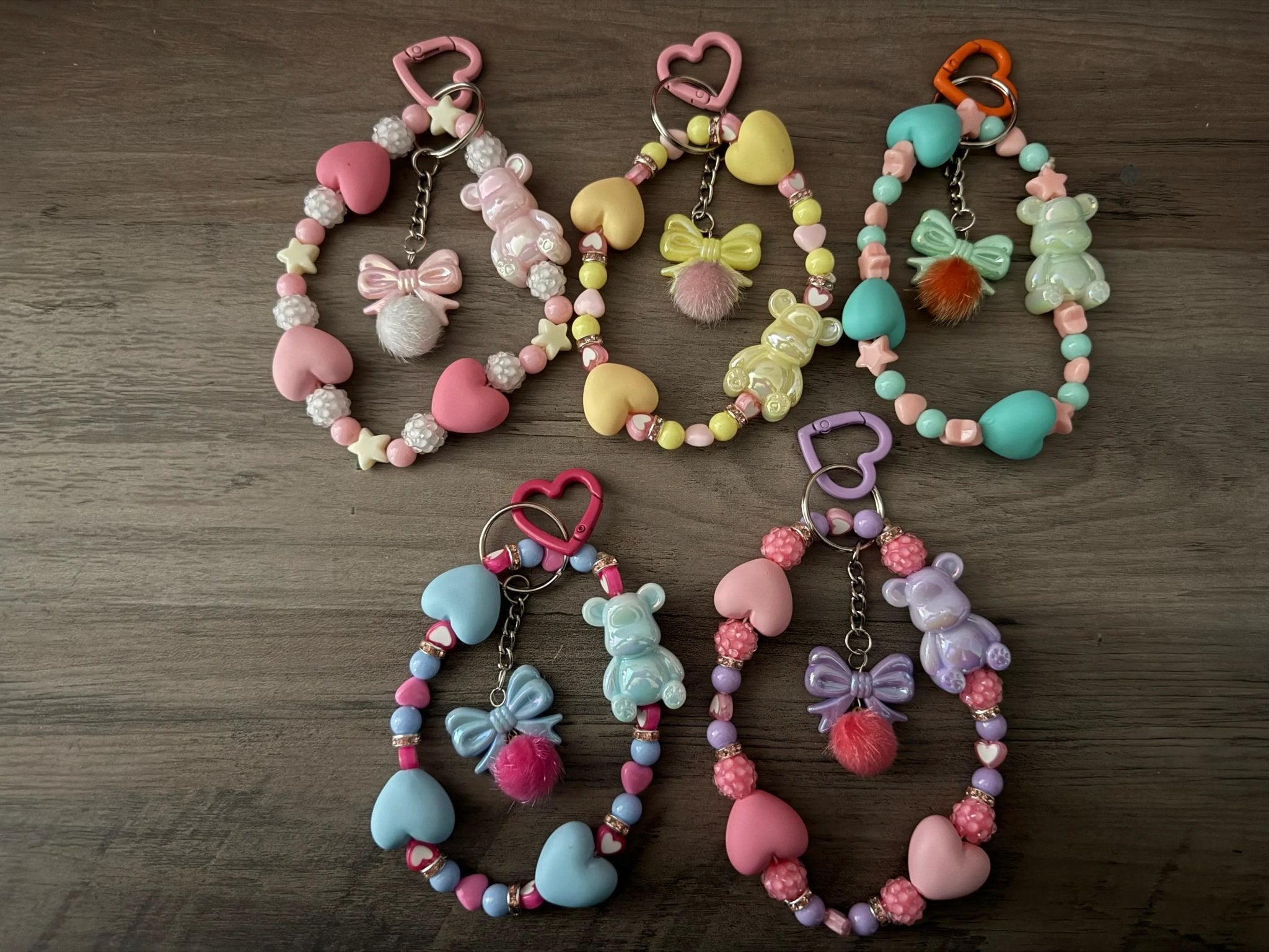Five colorful bag charms or phone charms with pom-poms, bear figures, bows, and heart beads.
