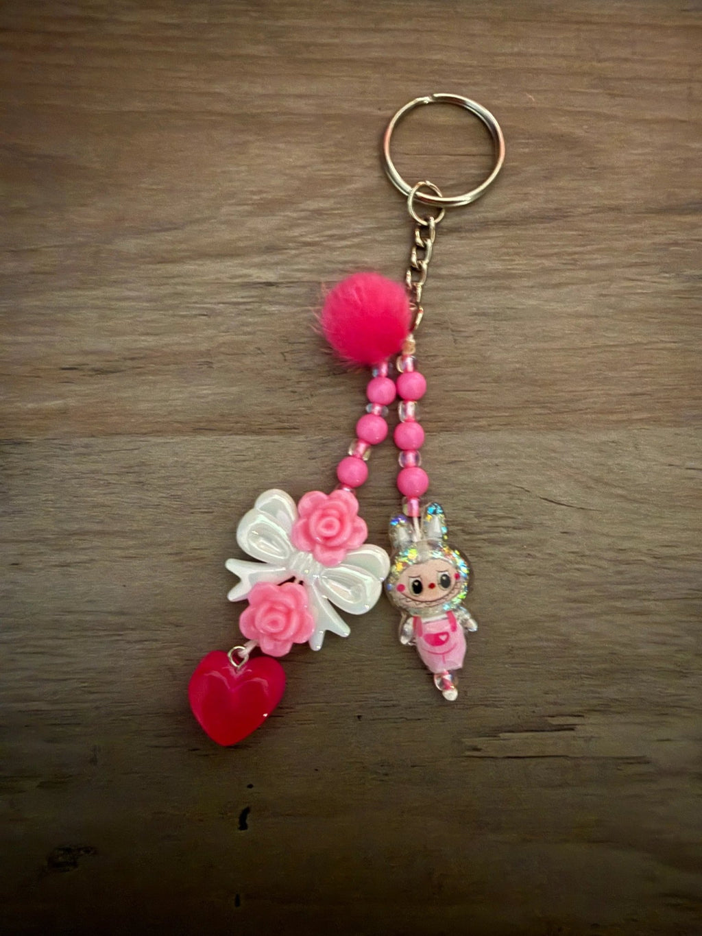 Pink keychain with a fluffy pom-pom, a bow with roses, a heart charm, and a cute character figure, strung with vibrant beads.