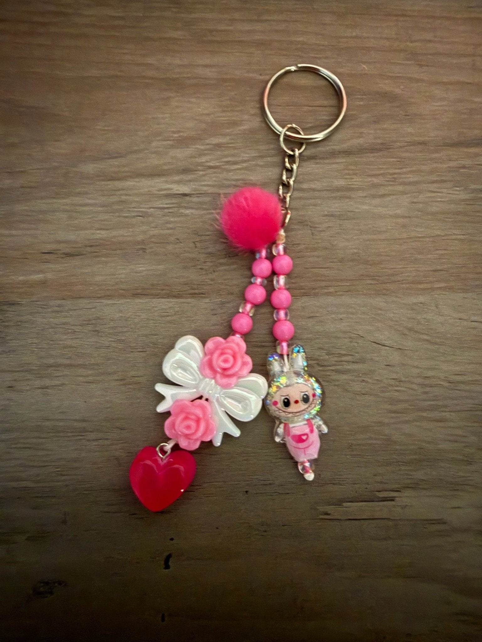 Pink keychain with a fluffy pom-pom, a bow with roses, a heart charm, and a cute character figure, strung with vibrant beads.